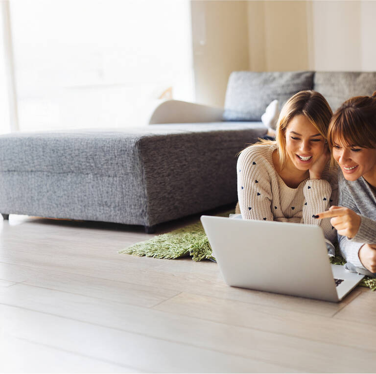 Zwei Frauen liegen entspannt vor einem Laptop und schauen gemeinsam darauf.