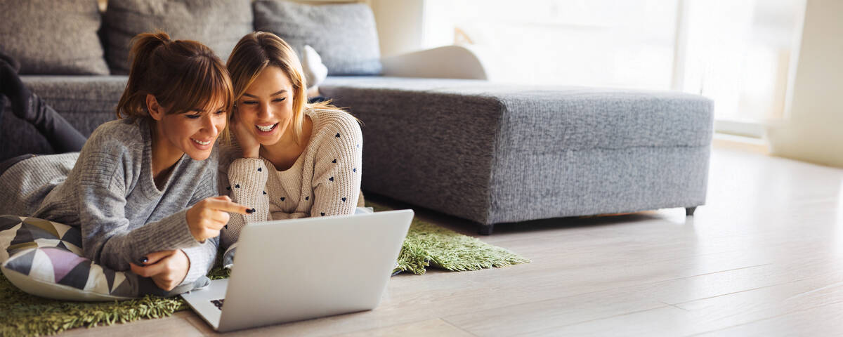 Zwei Frauen liegen entspannt vor einem Laptop und schauen gemeinsam darauf.