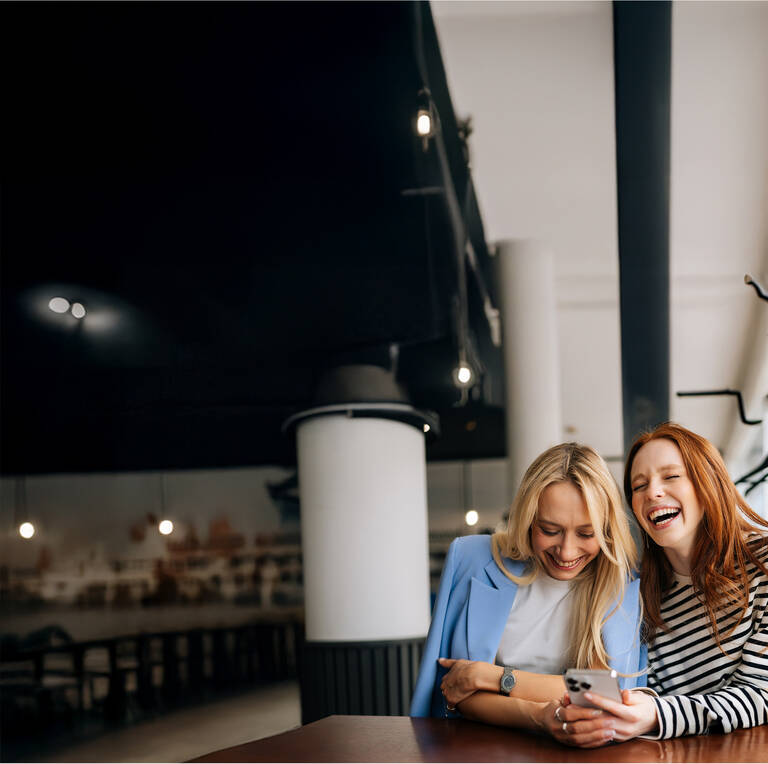Zwei Frauen sitzen in einem Café, lachen gemeinsam und schauen auf ihr Handy.