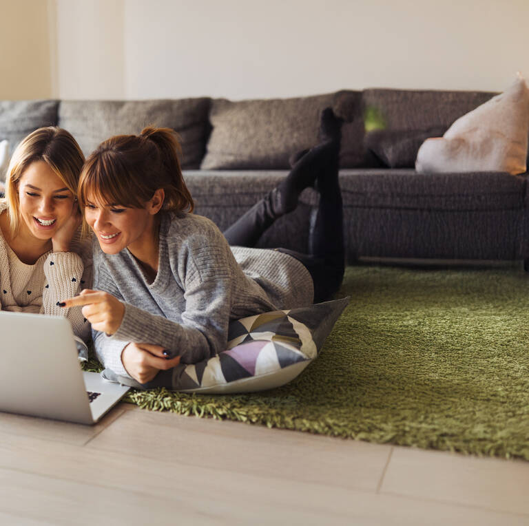 Zwei Frauen liegen entspannt vor einem Laptop und schauen gemeinsam darauf.
