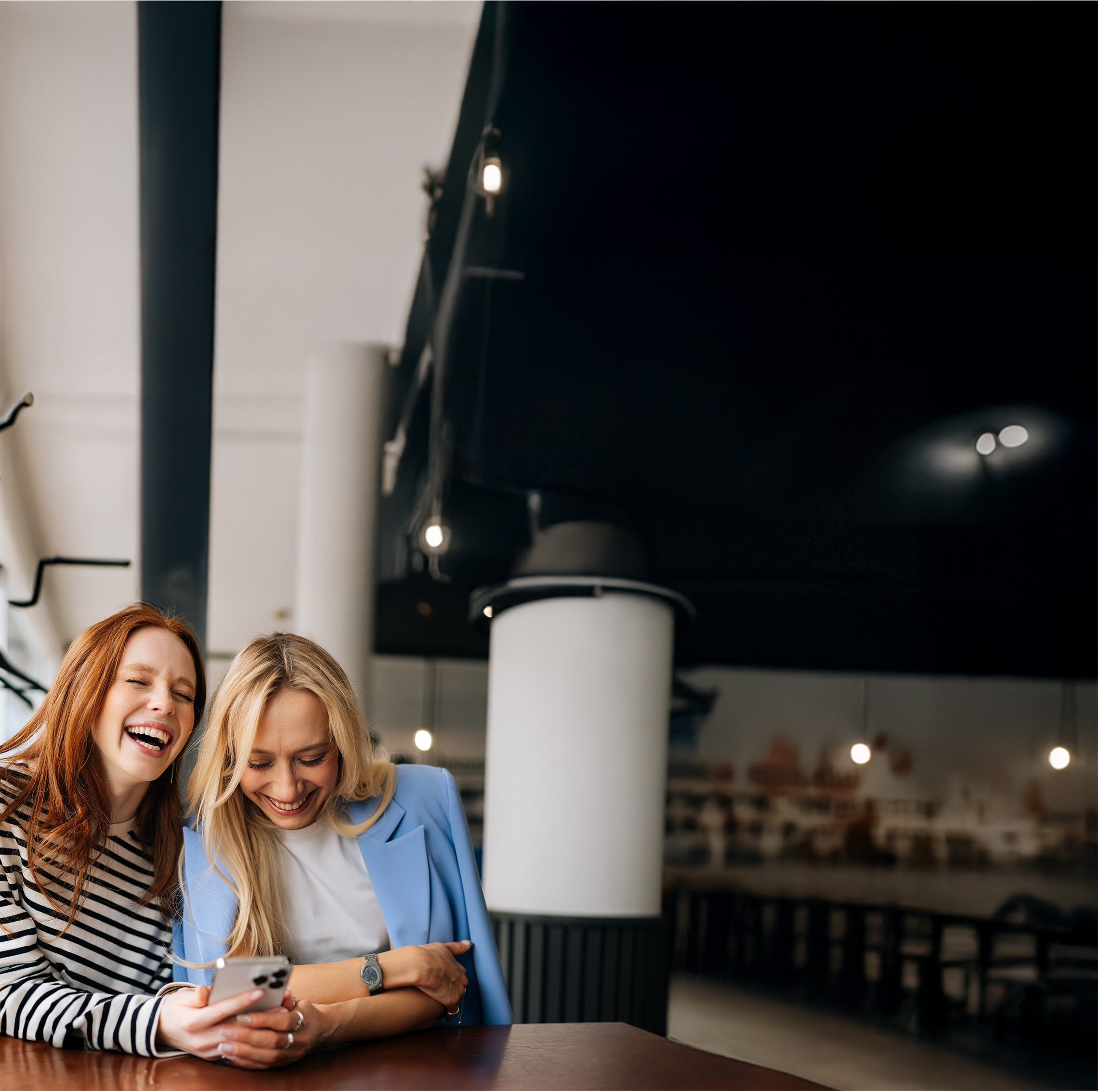 Gute Freundschaft zahlt sich aus! Zwei Frauen sitzen in einem Café, lachen gemeinsam und schauen auf ihr Handy.