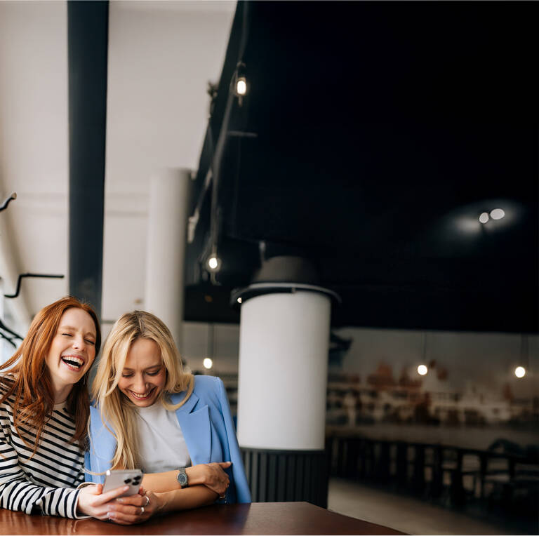 Zwei Frauen sitzen in einem Café, lachen gemeinsam und schauen auf ihr Handy.