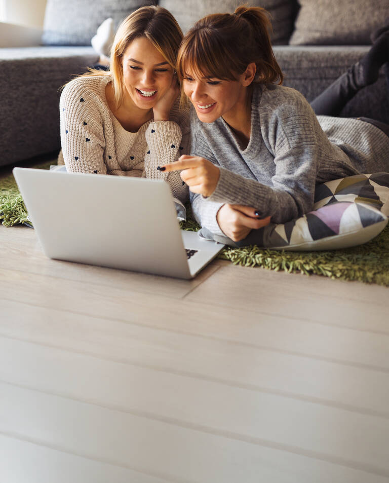 Zwei Frauen liegen entspannt vor einem Laptop und schauen gemeinsam darauf.