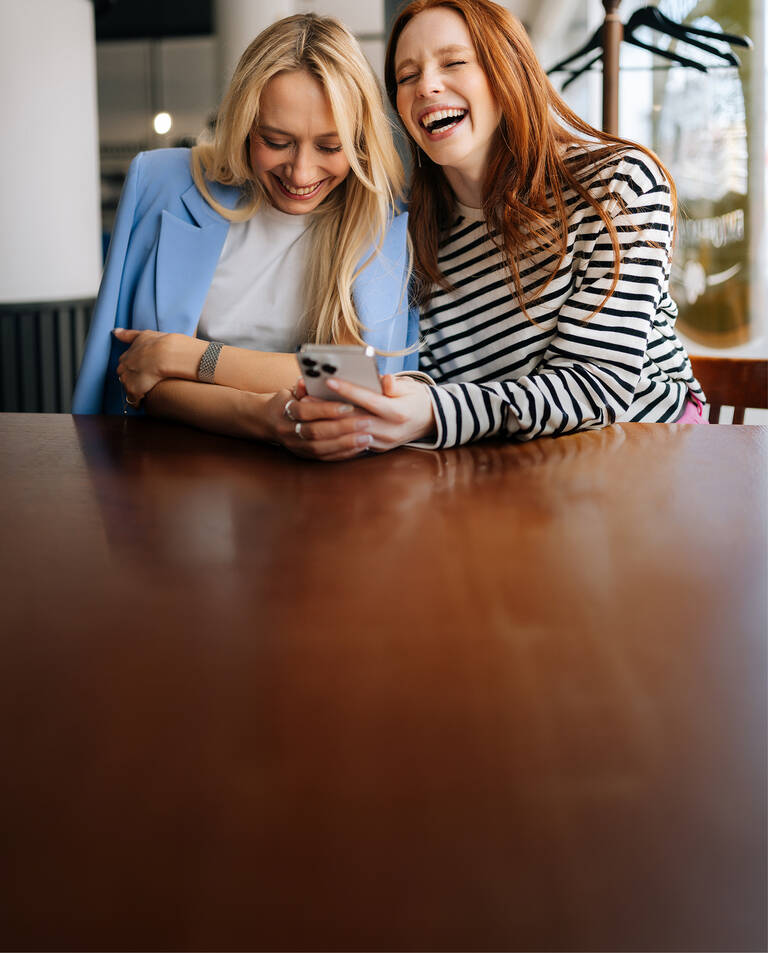 Zwei Frauen sitzen in einem Café, lachen gemeinsam und schauen auf ihr Handy.
