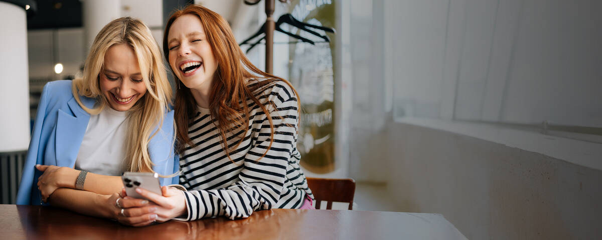 Zwei Frauen sitzen in einem Café, lachen gemeinsam und schauen auf ihr Handy.