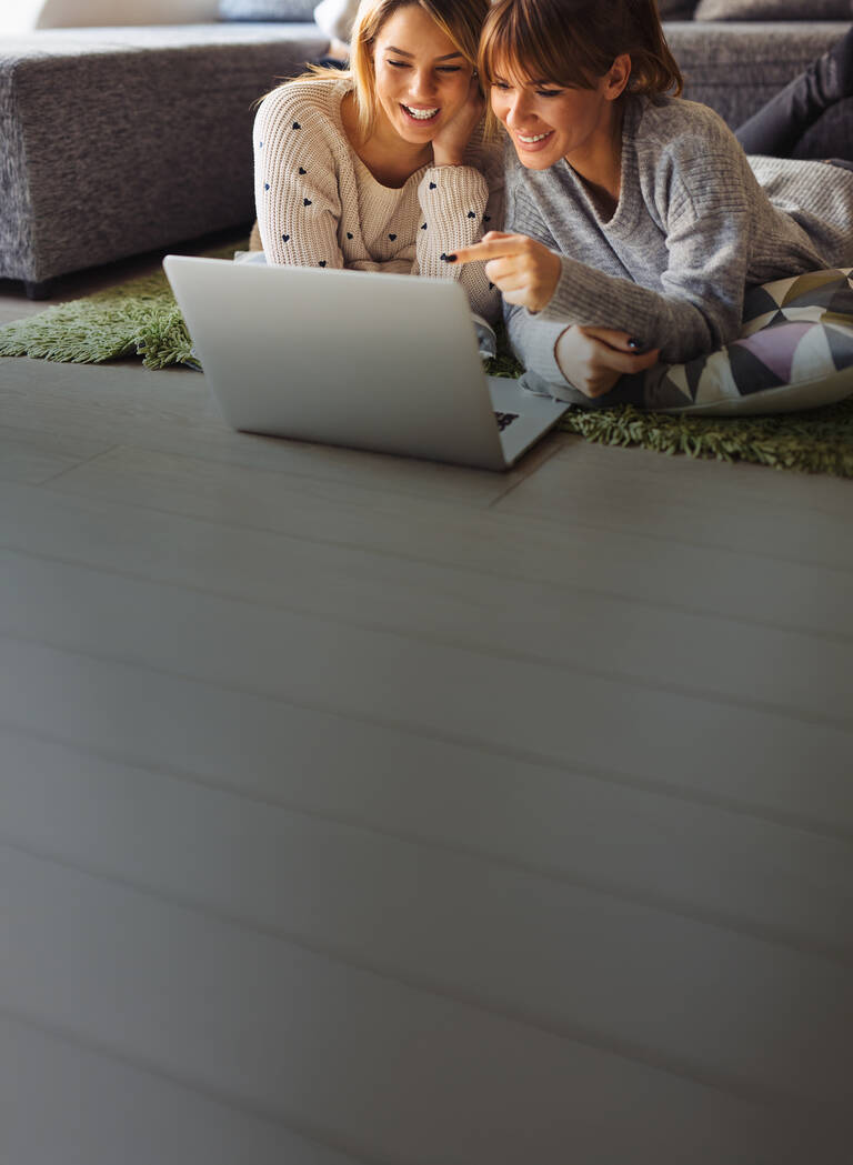 Zwei Frauen liegen entspannt vor einem Laptop und schauen gemeinsam darauf.