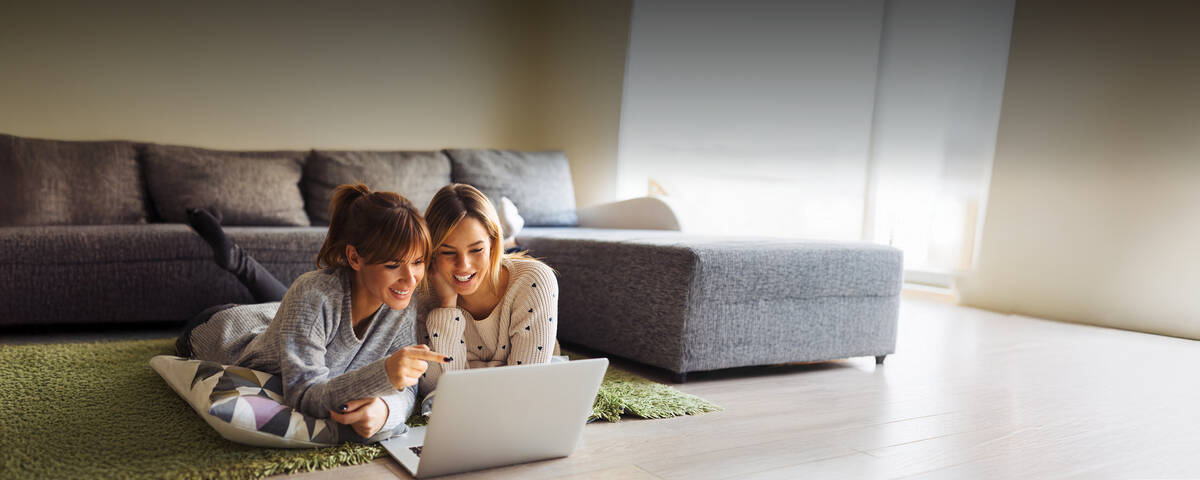 Zwei Frauen liegen entspannt vor einem Laptop und schauen gemeinsam darauf.