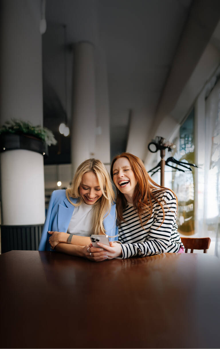 Zwei Frauen sitzen in einem Café, lachen gemeinsam und schauen auf ihr Handy.