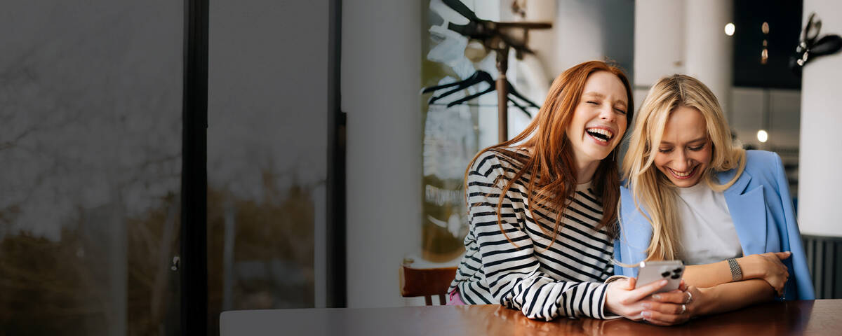 Zwei Frauen sitzen in einem Café, lachen gemeinsam und schauen auf ihr Handy.